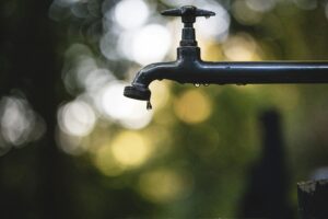 Saving Water At Home - Infinity Plumbing Services Close-up of a black outdoor faucet with droplets hanging from the spout against a bokeh background of green and white lights.
