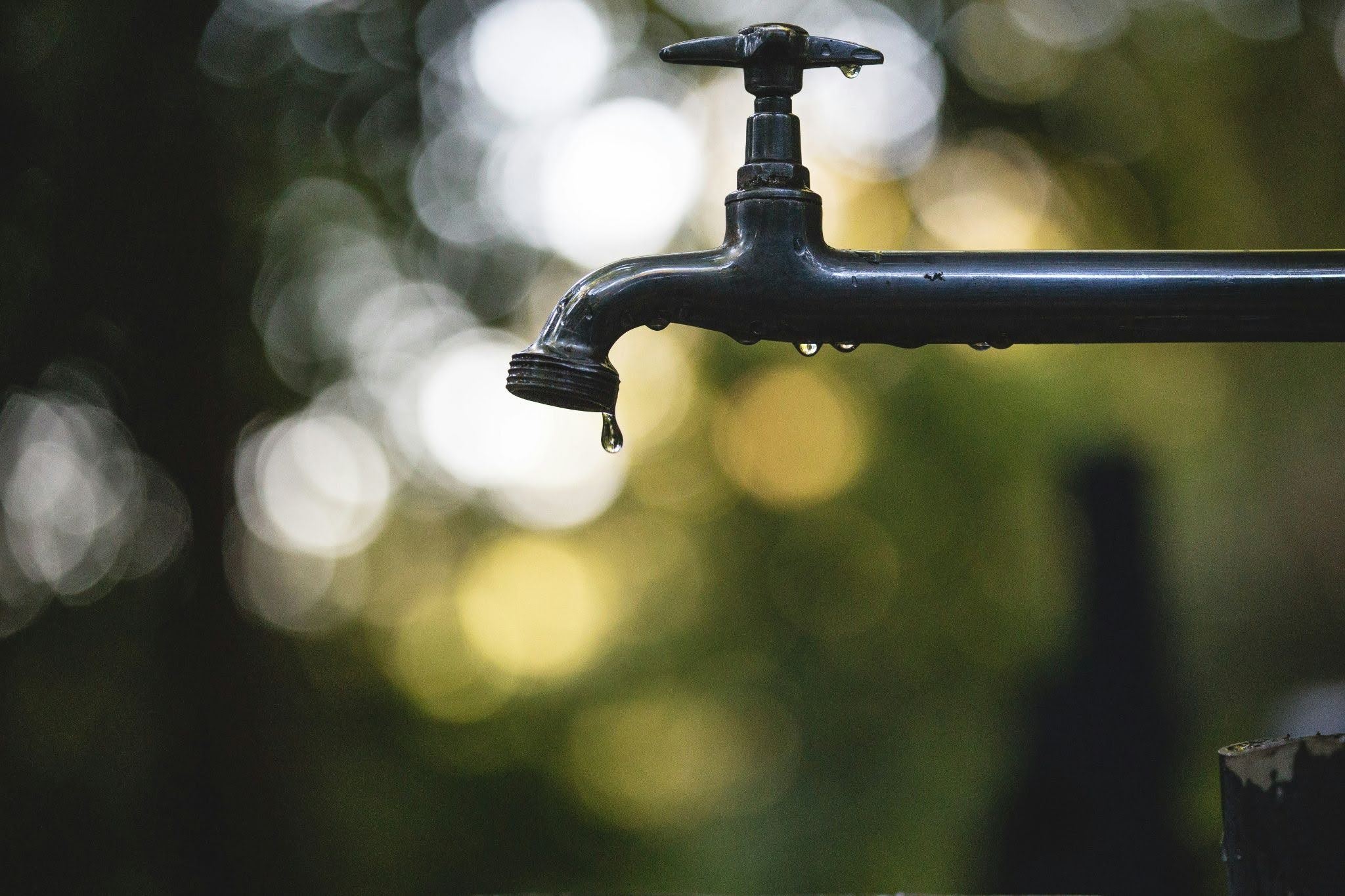 Saving Water At Home - Infinity Plumbing Services Close-up of a black outdoor faucet with droplets hanging from the spout against a bokeh background of green and white lights.