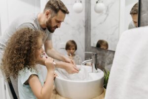 Kids brushing their teeth with their dad in the bathroom, smiling together during their morning routine in front of the sink. - Infinity Plumbing Services Kids brushing their teeth with their dad in the bathroom, smiling together during their morning routine in front of the sink.